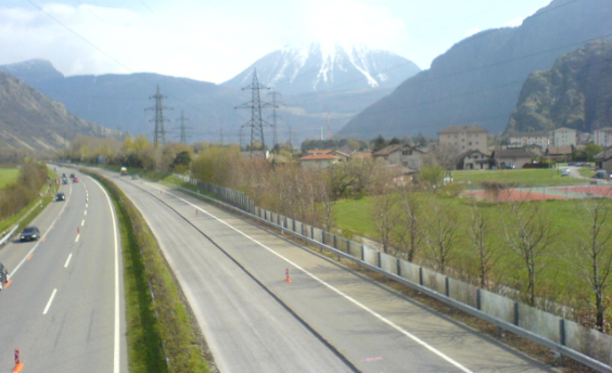 Vom unteren Bildrand nach oben links ist die vierspurige Autobahn mit einem Grünstreifen in der Mitte zu sehen. Rechts im Bild zieht sich eine Hochspannungsleitung durch, im Hintergrund sind Berge zu sehen.