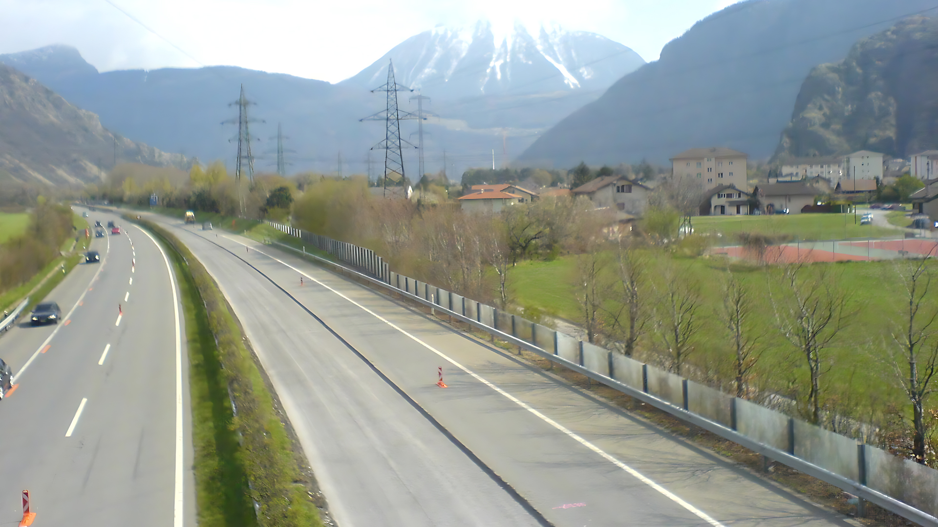 Vom unteren Bildrand nach oben links ist die vierspurige Autobahn mit einem Grünstreifen in der Mitte zu sehen. Rechts im Bild zieht sich eine Hochspannungsleitung durch, im Hintergrund sind Berge zu sehen.
